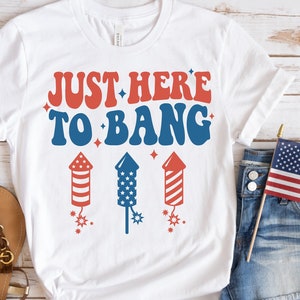 May include: White t-shirt with the text "Just Here To Bang" in red and blue letters with firework graphics. The shirt is on a white wooden background with a pair of brown sandals, denim shorts, and a small American flag.
