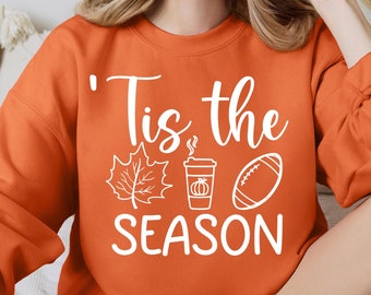 May include: A woman wearing an orange sweatshirt with the text "'Tis the season" and a graphic of a coffee cup, a leaf, and a football.