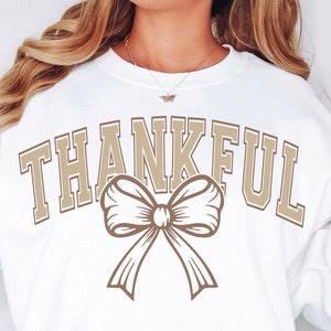 May include: A white sweatshirt with the word "THANKFUL" in tan block letters and a tan bow graphic. The sweatshirt has long sleeves and a crew neck. The overall design is simple and suitable for casual wear.