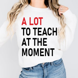 May include: White sweatshirt with the text "A LOT TO TEACH AT THE MOMENT" printed on the front. The word "LOT" is in red and the rest of the text is in black.