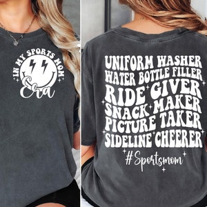 May include: A dark gray t-shirt with a white graphic on the back that says "Uniform Washer, Water Bottle Filler, Ride Giver, Snack Maker, Picture Taker, Sideline Cheerer #Sportsmom". The front of the shirt has a white graphic of a smiley face with lightning bolts and the text "I'm My Sports Mom Era".