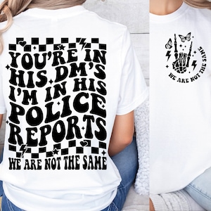 May include: White t-shirt with black text that reads "You're in his DM's I'm in his police reports We are not the same". The text is in a retro font style. The back of the shirt has a checkered pattern. The front of the shirt has a black graphic of a skeleton hand with lightning bolts and butterflies.