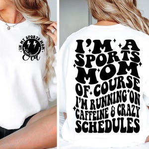 Pode incluir: Camisola branca com texto preto que diz "Eu sou uma m&atilde;e esportiva, claro que estou correndo com cafe&iacute;na e hor&aacute;rios loucos". O texto est&aacute; em um estilo de fonte retr&ocirc;. H&aacute; um design menor na frente da camiseta que diz "Na minha era de m&atilde;e esportiva" com um rosto sorridente e raios.