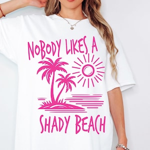 May include: White t-shirt with a pink graphic design that says "Nobody likes a shady beach". The graphic includes palm trees, a sun, and a beach.