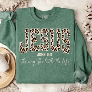 May include: A teal sweatshirt featuring the word "JESUS" in large leopard print lettering. The text "JOHN 14:6" and the phrase "the way the truth the life" are below the word "JESUS" in a cursive font.