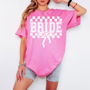 Puede incluir: Una camiseta rosa con la palabra "BRIDE" en letras blancas, sobre un patr&oacute;n de cuadros. Una cinta blanca est&aacute; debajo de la palabra. La camiseta es de corte holgado.
