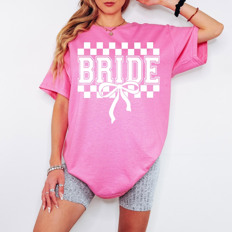 Puede incluir: Una camiseta rosa con la palabra "BRIDE" en letras blancas, sobre un patr&oacute;n de cuadros. Una cinta blanca est&aacute; debajo de la palabra. La camiseta es de corte holgado.