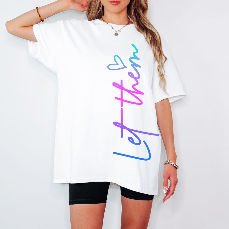 May include: White t-shirt with the text "Let them" in a gradient of blue, purple, and pink. The shirt has a relaxed fit and is paired with black shorts. The text includes a small heart symbol.