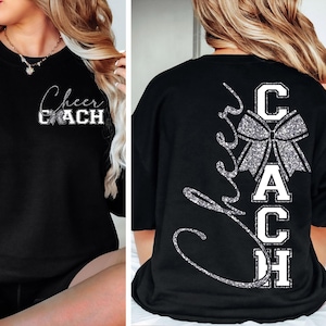 Puede incluir: Sudadera negra con la inscripción "Cheer Coach" en blanco y purpurina plateada. El diseño frontal presenta el texto y un lazo. La parte trasera de la camiseta tiene la palabra "Cheer" en cursiva y la palabra "Coach" en letras de bloque con un lazo.