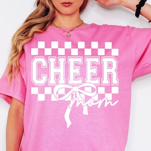 May include: Pink t-shirt with a white checkered pattern, the words "CHEER MOM" and a white bow.
