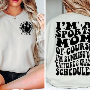 Pode incluir: Uma camisa de moletom branca com texto preto que diz "Eu sou uma m&atilde;e esportiva, claro que estou correndo com cafe&iacute;na e hor&aacute;rios loucos". A frente da camisa de moletom tem um rosto sorridente preto com raios e o texto "Na minha era de m&atilde;e esportiva".