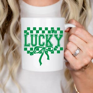 Pu&ograve; includere: Una tazza in ceramica bianca con un motivo a scacchi verdi e un fiocco verde. La parola "LUCKY" &egrave; scritta in lettere verdi sulla tazza.