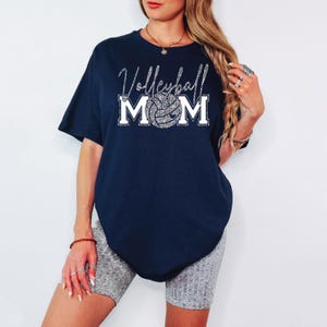 May include: Navy blue t-shirt with the text "Volleyball Mom" in a silver glitter font. The word "Mom" is above a volleyball graphic. The shirt is a short-sleeved, oversized fit.