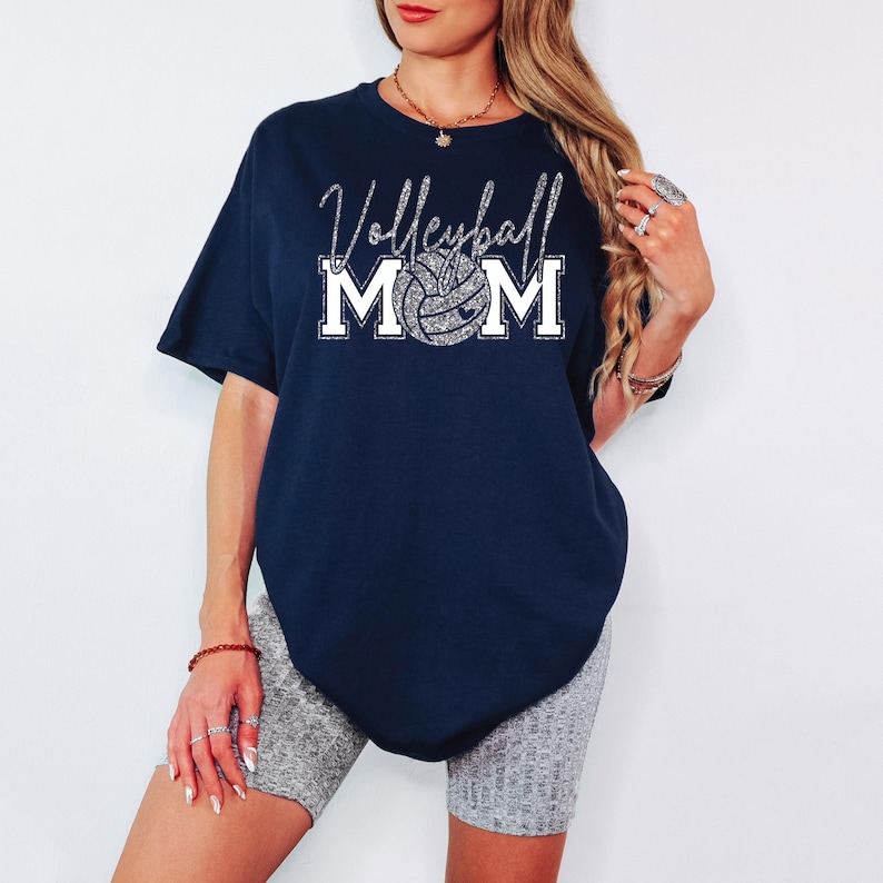 May include: Navy blue t-shirt with the text "Volleyball Mom" in a silver glitter font. The word "Mom" is above a volleyball graphic. The shirt is a short-sleeved, oversized fit.
