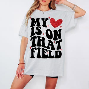 Pu&ograve; includere: Una maglietta bianca con una grafica nera che dice "My heart is on that field" con un cuore rosso glitterato.