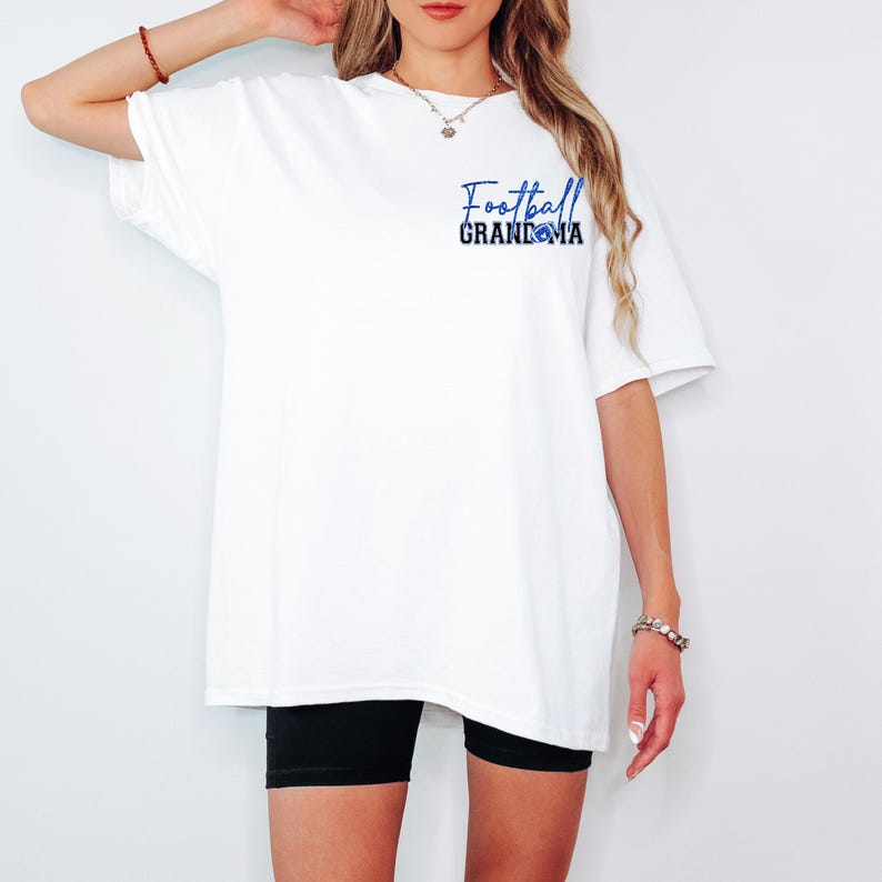 Pu&ograve; includere: T-shirt bianco con la scritta "Football Grandma" in blu e una grafica di football americano. La maglietta &egrave; di taglio ampio e indossata con pantaloncini neri. Lo sfondo &egrave; bianco.