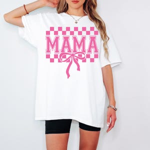 May include: White oversized t-shirt with a pink checkered pattern and the word "MAMA" in pink block letters. A pink bow is centered below the text.