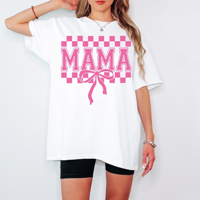 May include: White oversized t-shirt with a pink checkered pattern and the word "MAMA" in pink block letters. A pink bow is centered below the text.