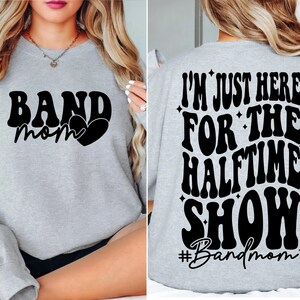 Puede incluir: Sudadera gris con el texto "Band Mom" y un coraz&oacute;n en la parte delantera. La parte posterior de la sudadera tiene el texto "I'm just here for the halftime show #Bandmom" en una fuente retro.