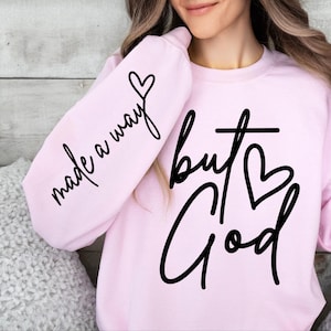 May include: Pink sweatshirt with black text that reads "made a way but God" with a heart symbol on the sleeve and a cross symbol on the front.