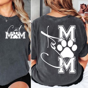 May include: Dark gray t-shirt with the words "Cat Mom" in a white script and block letters, with a paw print in the center. The back of the shirt also features the words "Cat Mom" in a similar design.