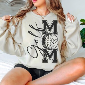 Może przedstawiać: Kremowa bluza z napisem "Golf Mom" w czarnym, ozdobnym pismem i grafiką piłki golfowej. Słowo "Mom" jest dużymi literami.