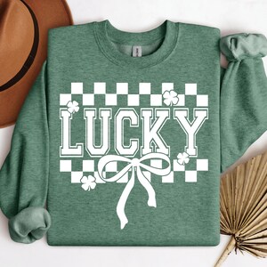 Pu&ograve; includere: Felpa verde con un motivo a scacchi bianchi, un fiocco bianco e la parola "LUCKY" in lettere bianche.
