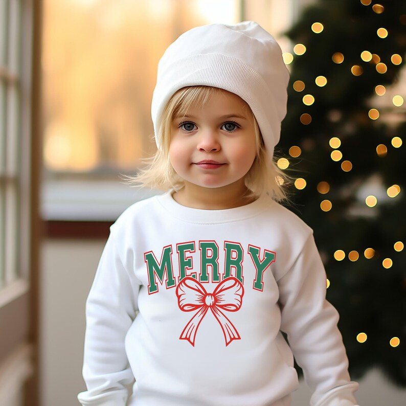 May include: A white sweatshirt with a green and red "Merry" graphic featuring a red bow. The sweatshirt is being worn by a young child wearing a white beanie.