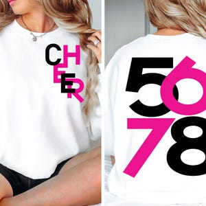 May include: White sweatshirt with black and pink text. The front of the sweatshirt says "CHER" in black letters with a pink outline. The back of the sweatshirt says "5678" with the numbers 5 and 7 in pink and the numbers 6 and 8 in black.
