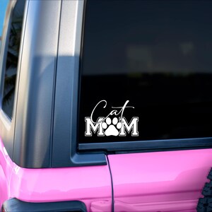 May include: A white vinyl decal on a pink vehicle window. The decal reads "Cat Mom" with a paw print in the center. The vehicle is parked on a sandy beach with palm trees in the background.
