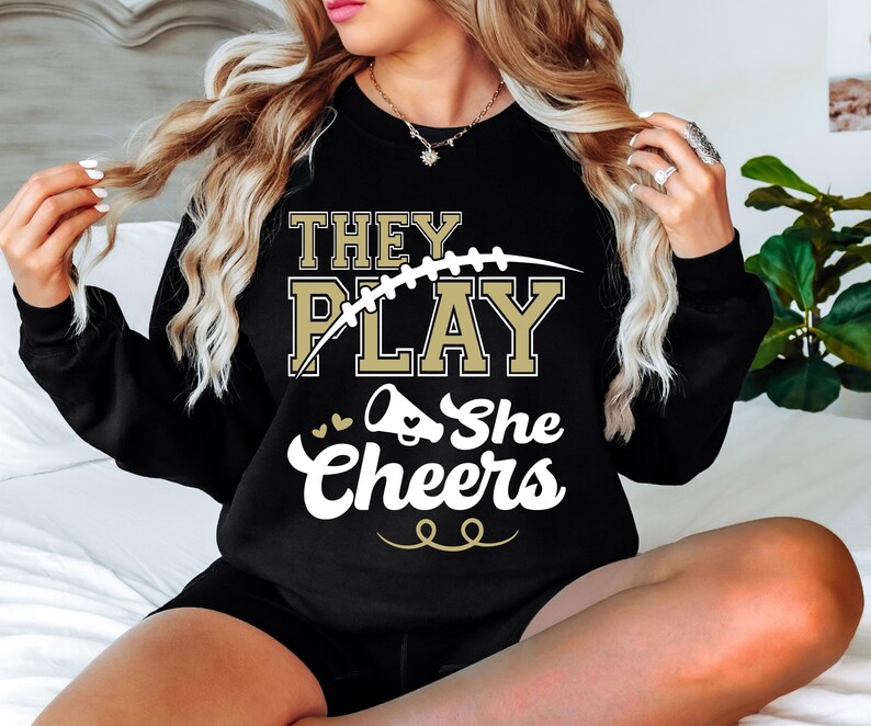 May include: Black sweatshirt with gold lettering that reads "They Play She Cheers" with a football and a cheerleader megaphone graphic.