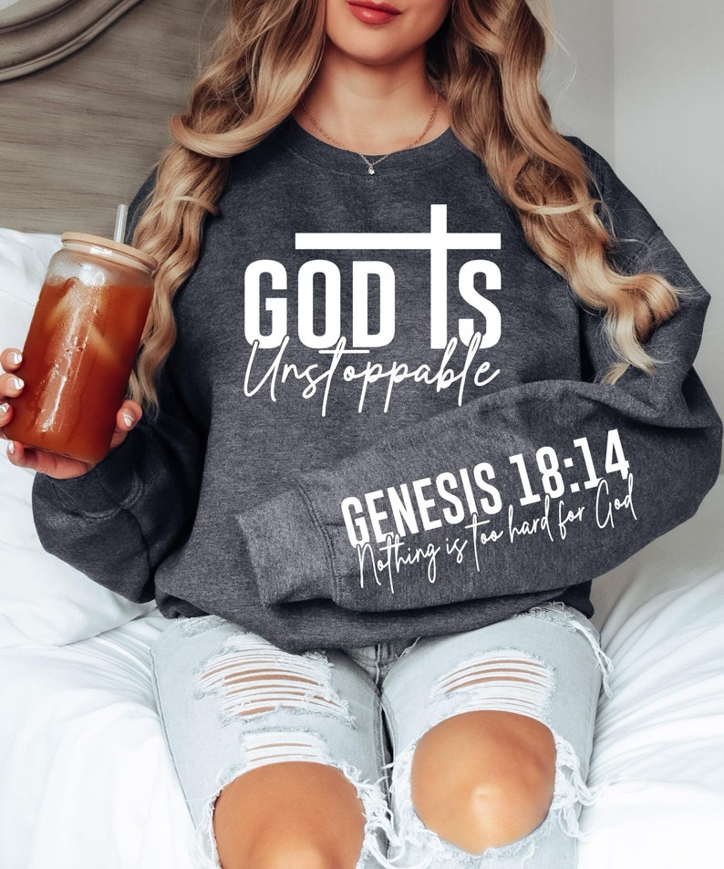 May include: Dark gray sweatshirt with the text "GOD IS Unstoppable" and a cross graphic. The sleeve features the text "GENESIS 18:14 Nothing is too hard for God" in white. The sweatshirt is worn with light wash jeans.