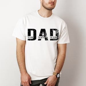 K&ouml;nnte beinhalten: Ein wei&szlig;es T-Shirt mit kurzen &Auml;rmeln und dem schwarzen, im Used-Look gestalteten Schriftzug DAD in gro&szlig;en Druckbuchstaben. Unter jedem Buchstaben steht in kleinerer schwarzer Schrift THE MAN, THE MYTH und THE LEGEND als Grafik.