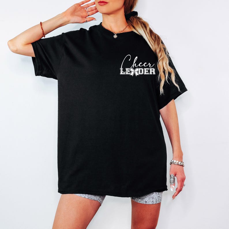 Puede incluir: Camiseta negra de gran tama&ntilde;o con texto blanco que dice "Cheer Leader" y un gr&aacute;fico de lazo blanco.