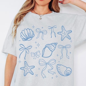 Può includere: T-shirt grigio chiaro con un design blu a conchiglie, fiocchi e stelle marine. La maglietta ha un girocollo e maniche corte. Il design include diversi fiocchi, conchiglie, stelle marine e accenti di stelle scintillanti.