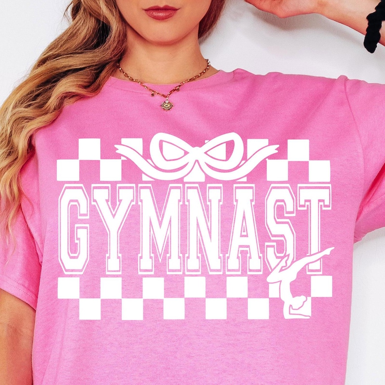 May include: Pink t-shirt with a white checkered pattern, a white ribbon, the word "GYMNAST" in white block letters, and a white silhouette of a gymnast.