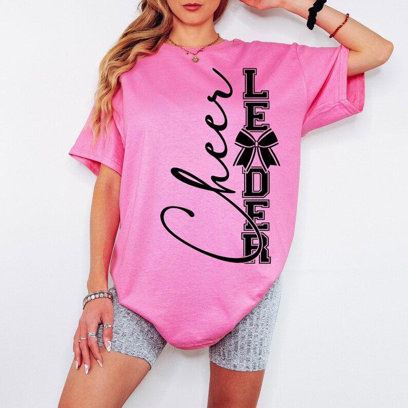 Puede incluir: Camiseta rosa con texto negro que dice "Cheer Leader" con un lazo negro en el medio de la palabra "Leader".