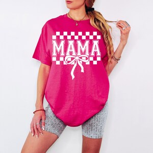 May include: A pink t-shirt with a white checkered pattern and the word "MAMA" in white letters. A white bow is centered below the word.