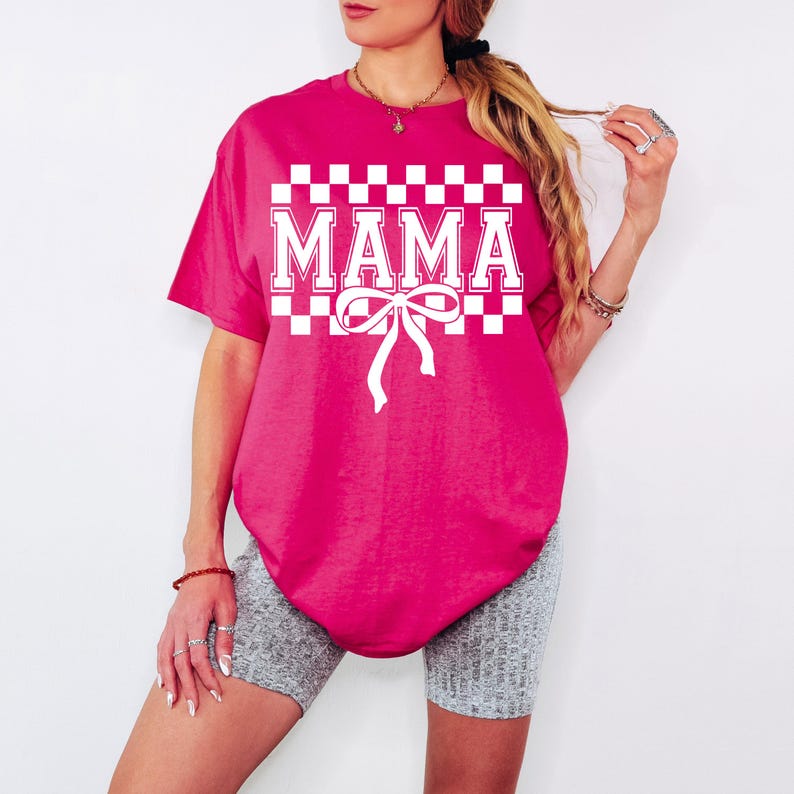 May include: A pink t-shirt with a white checkered pattern and the word "MAMA" in white letters. A white bow is centered below the word.