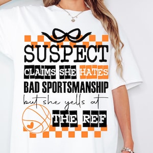 May include: White t-shirt with a black and orange checkered pattern and the text "SUSPECT CLAIMS SHE HATES BAD SPORTSMANSHIP but she yells at THE REF" with a basketball graphic.