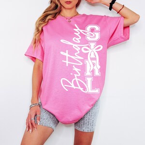 May include: A bright pink t-shirt featuring the words "Birthday Girl" in white, stylish script and block letters. The shirt has short sleeves and a loose fit. The design includes a bow, making it a fun, celebratory item.