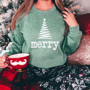 May include: A green sweatshirt with a white Christmas tree design and the word "merry" printed below it. The person is holding a red mug with a white mustache design. The person is wearing black pants with white snowflake patterns.