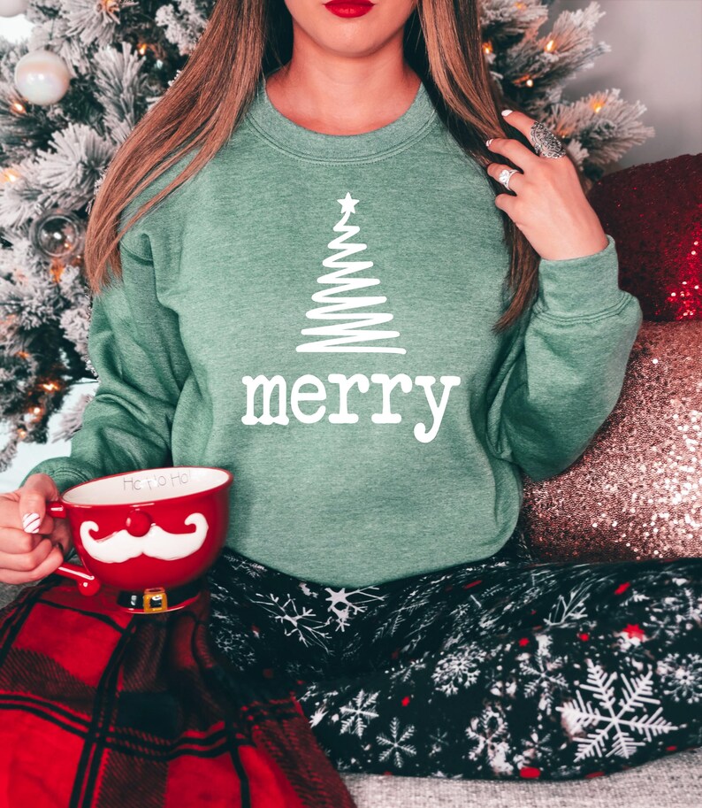 May include: A green sweatshirt with a white Christmas tree design and the word "merry" printed below it. The person is holding a red mug with a white mustache design. The person is wearing black pants with white snowflake patterns.