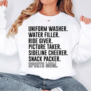 May include: White sweatshirt with black text that reads "Uniform Washer. Water Filler. Ride Giver. Picture Taker. Sideline Cheerer. Snack Packer. Sports Mom."