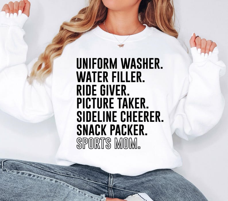 May include: White sweatshirt with black text that reads "Uniform Washer. Water Filler. Ride Giver. Picture Taker. Sideline Cheerer. Snack Packer. Sports Mom."