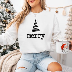 May include: White sweatshirt with a black Christmas tree design and the word "merry" in black. The person is holding a white mug with a Santa Claus illustration. The background includes a decorated Christmas tree.