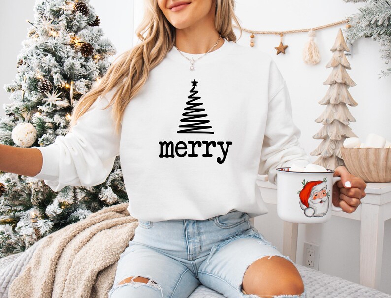 May include: White sweatshirt with a black Christmas tree design and the word "merry" in black. The person is holding a white mug with a Santa Claus illustration. The background includes a decorated Christmas tree.