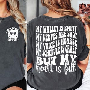 Könnte beinhalten: Ein dunkelgraues T-Shirt mit einem weißen Smiley mit Herzen als Augen und dem Text "Dance Mom" auf der Vorderseite. Die Rückseite des Shirts hat weißen Text, der lautet: "Mein Portemonnaie ist leer, meine Nerven sind blank, meine Stimme ist heiser, mein Zeitplan ist verrückt, aber mein Herz ist voll."