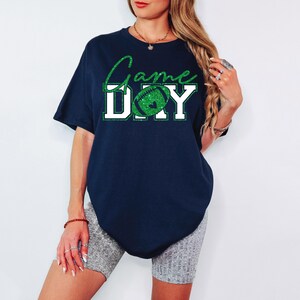 K&ouml;nnte beinhalten: Marineblaues T-Shirt mit dem Text "Game Day" in wei&szlig;en Blockbuchstaben und einer gr&uuml;nen Glitzer-Football-Grafik. Das Shirt hat eine lockere Passform und ist f&uuml;r den Freizeitgebrauch geeignet.