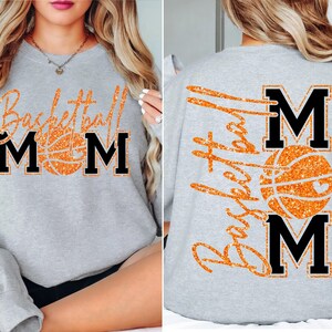 May include: A light gray sweatshirt with the words "Basketball Mom" in orange glitter lettering and a basketball graphic. The letters "MOM" are in black block letters. The back of the sweatshirt also features the words "Basketball Mom".